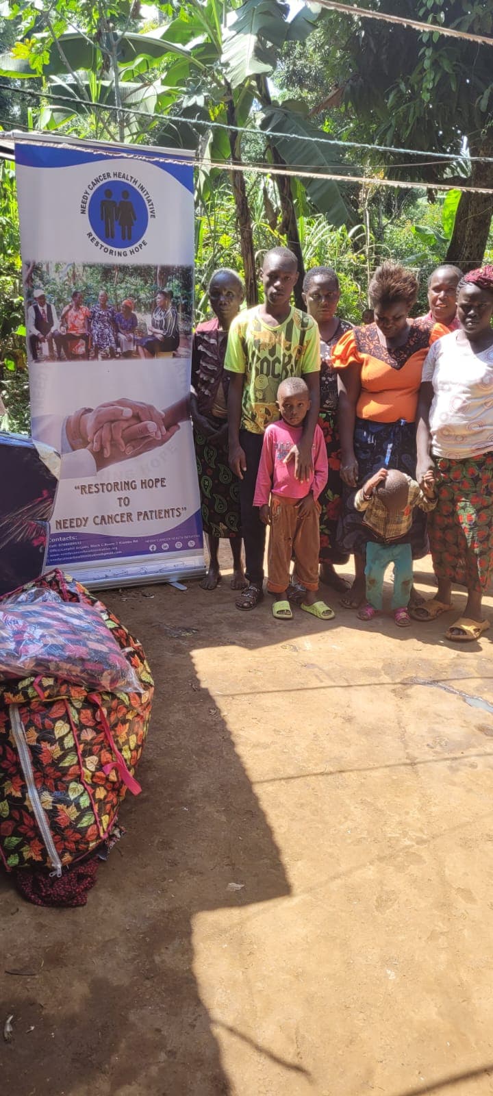 Families standing beside the NCHI “Restoring Hope” banner during a home visit, with bags of supplies in the foreground